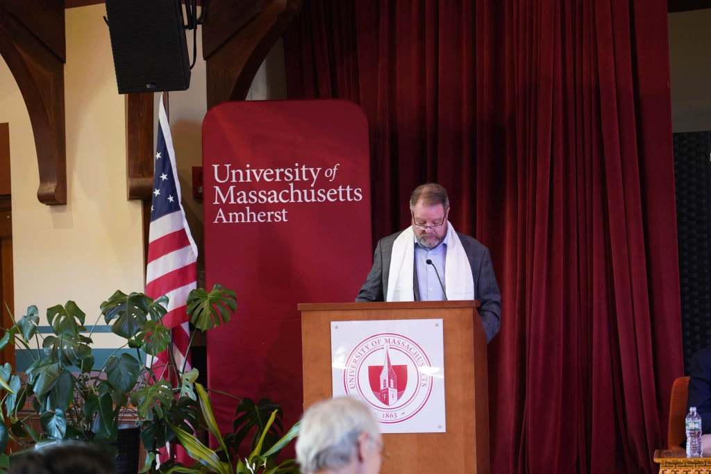 A speaker addressing an audience at the University of Massachusetts Amherst, with an American flag and a red backdrop featuring the university logo.
