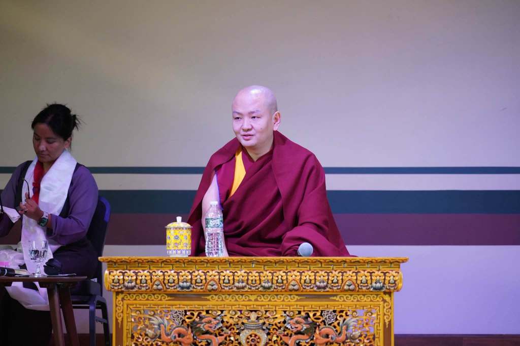 A monk in traditional attire sitting at a decorated table, engaging with an audience. A woman in the background prepares materials.