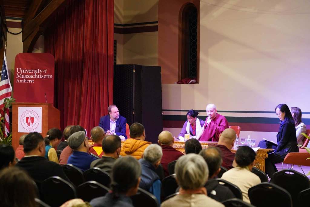 A panel discussion taking place at the University of Massachusetts Amherst, featuring speakers on stage, including a monk, with an audience of engaged attendees.
