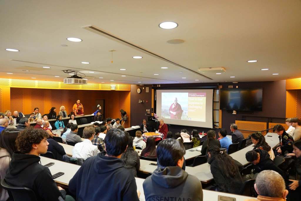 A large group of people seated in a classroom for a lecture, with an image of a monk displayed on a screen, discussing themes of compassion and challenging times.