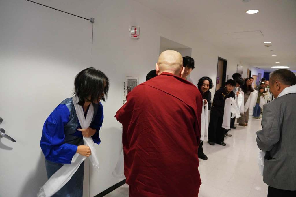 A group of individuals in a hallway, including a monk in traditional robes and people dressed in various cultural attire, participating in a ceremony while holding white scarves.