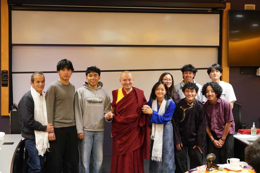 A group of nine individuals posing for a photo, including a monk in traditional robes, in a classroom or conference setting.