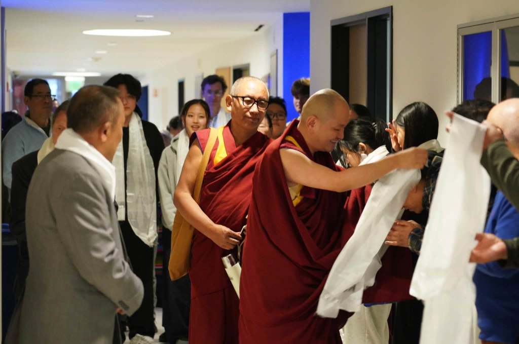 Buddhist monks in traditional robes interacting with a crowd, with people receiving ceremonial scarves in a hallway.