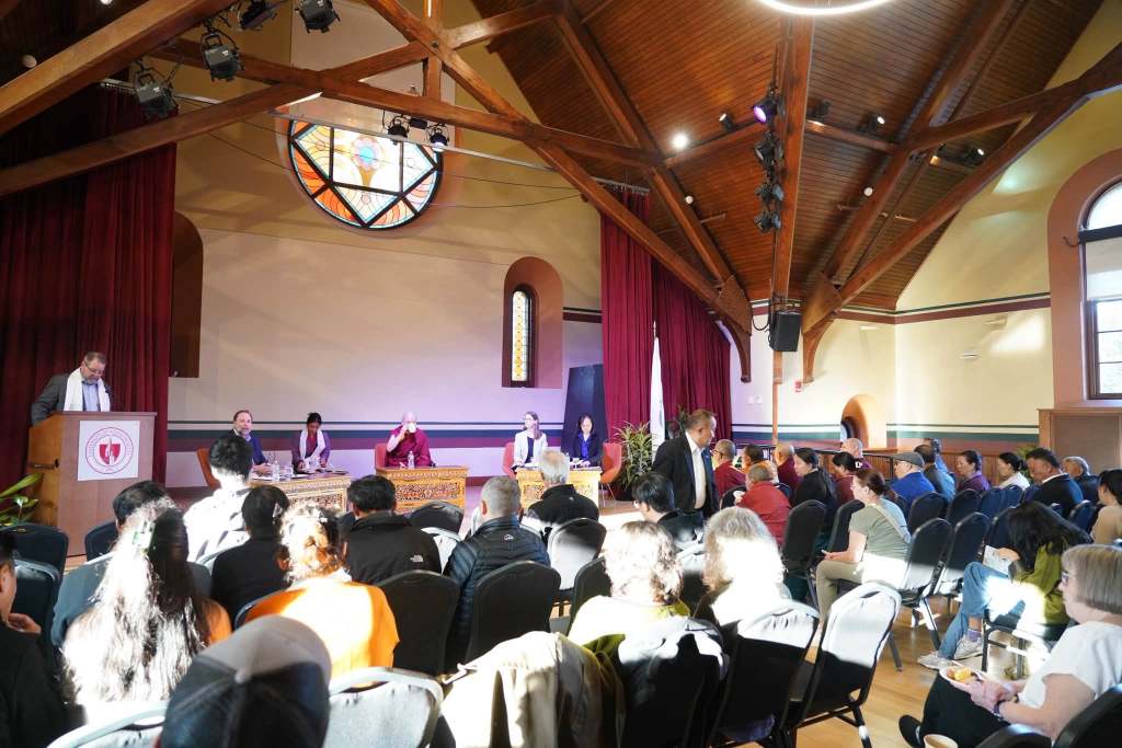 A panel discussion taking place in a hall with wooden beams and stained glass windows. Several speakers are seated at a table, addressing an audience of diverse individuals seated in rows.