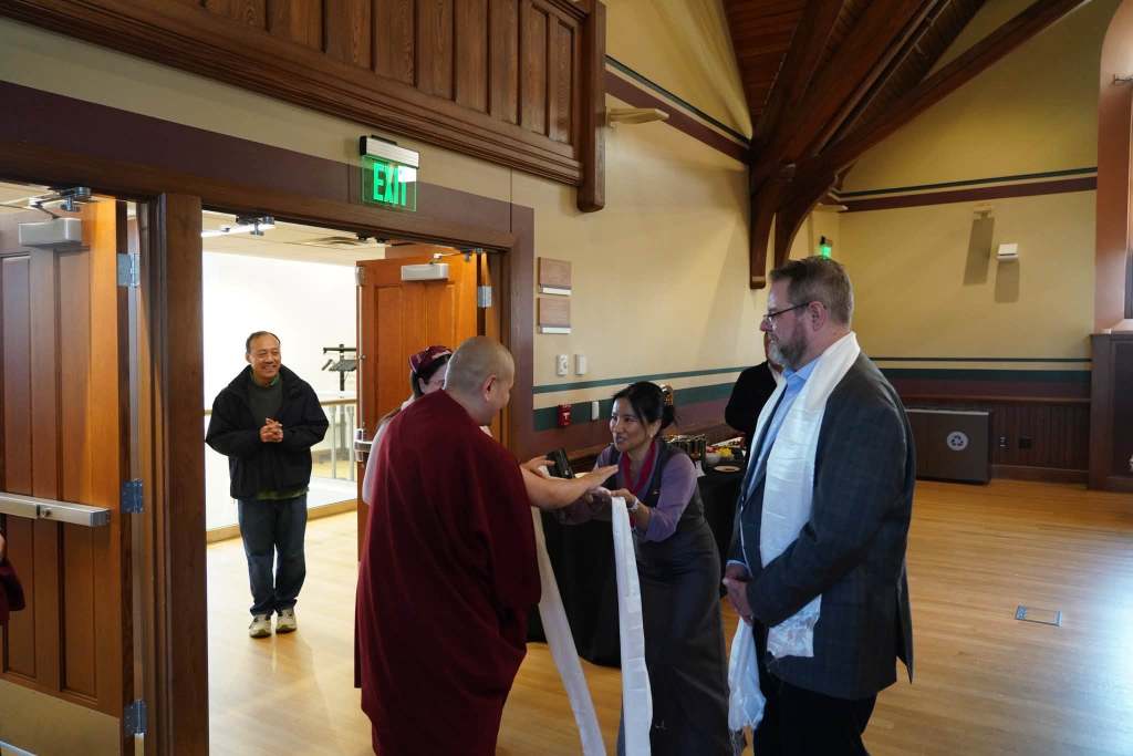 A Buddhist monk presenting a ceremonial scarf to two individuals in a warm, wooden interior space, while another person looks on with a smile.